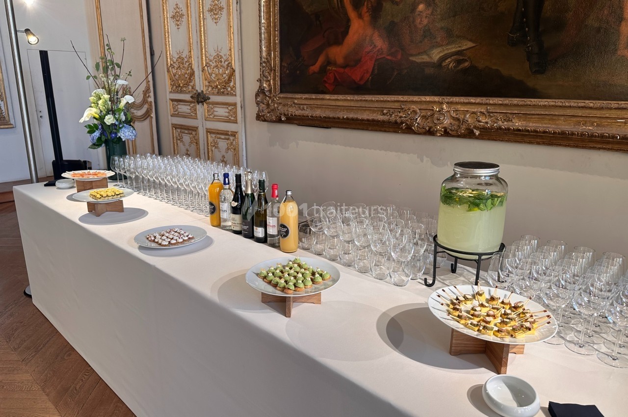 Traiteur Arômes et Sens Sèvres #1 Buffet élégant avec amuse-bouches, boissons variées et décoration florale dans une salle ornée de tableaux et moulures…