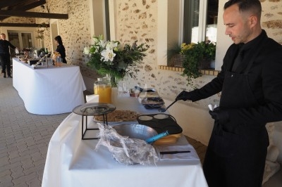 Un cuisinier prépare des crêpes sur une plaque chauffante lors d'un buffet en extérieur près d'un bâtiment en pierre.