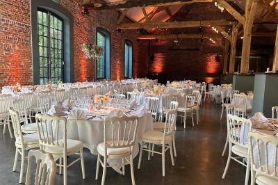 Salle de réception avec tables rondes dressées, chaises blanches et éclairage chaleureux dans un bâtiment en briques.
