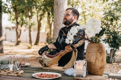 Traiteur Stéphane Portier Traiteur Le Plessis-Macé #1 Un homme découpe un jambon cru sur une table en bois en extérieur, entouré d'arbres et de vaisselle.