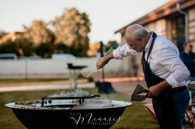 Traiteur Au Bois du Bon Sejour Aubeterre #2 Un homme en tablier cuisine sur un brasero en plein air, saupoudrant des ingrédients sur une plaque chauffante.