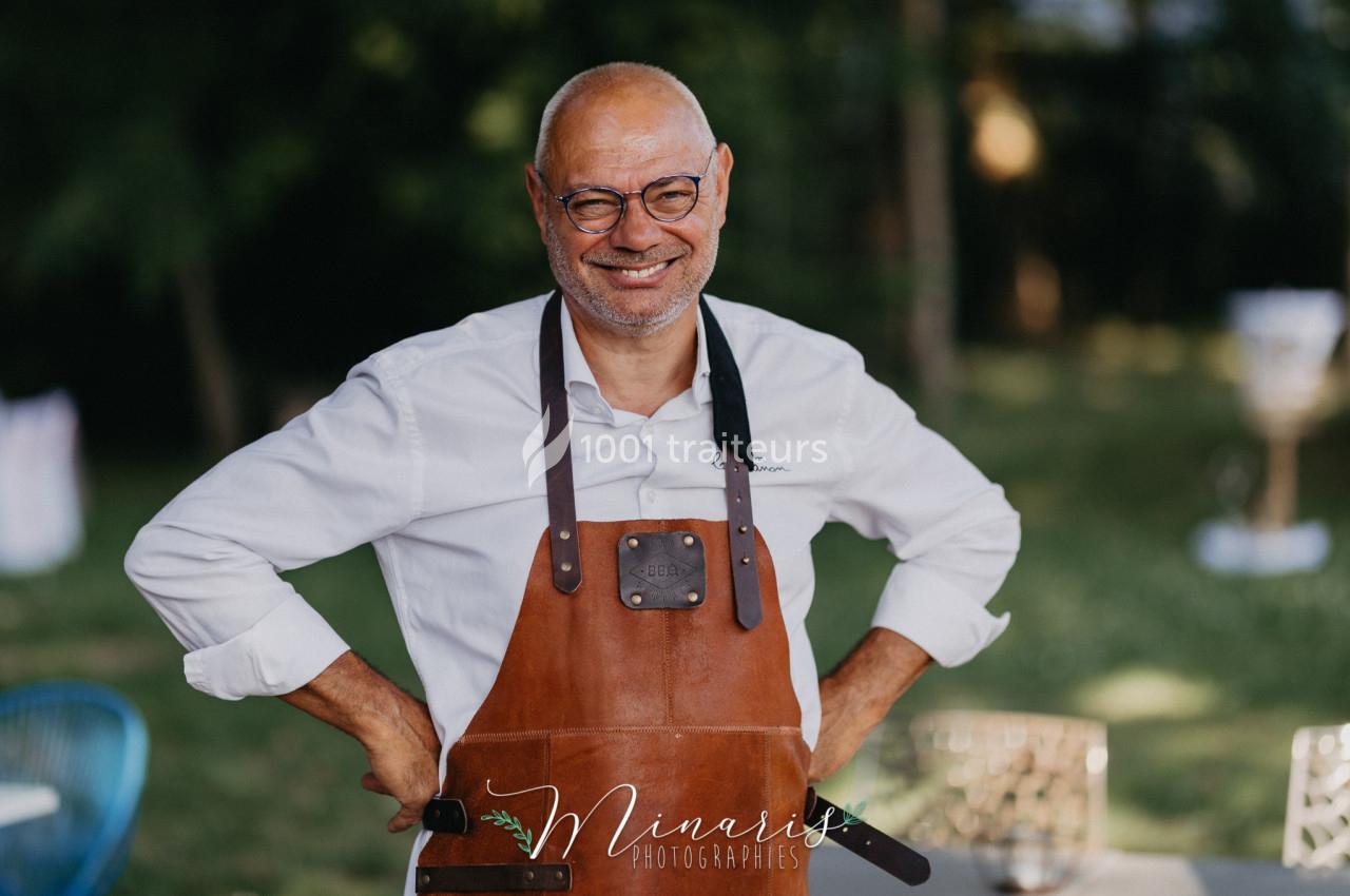 Traiteur Au Bois du Bon Sejour Aubeterre #1 Homme souriant portant un tablier en cuir, debout en extérieur avec un fond flou de verdure et de mobilier.