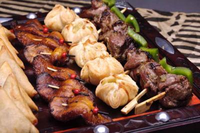 Assortiment de brochettes de viande grillée, beignets et samoussas disposés sur un plateau en bois.