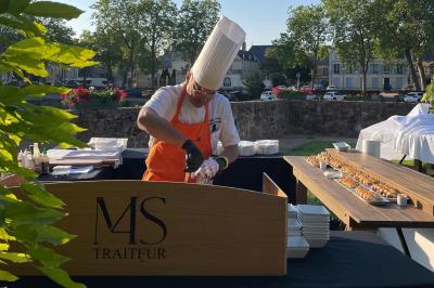 Traiteur Le Moulin des Quatre Saisons La Flèche #1 Un chef en tenue blanche et tablier orange prépare un plat derrière un comptoir en bois marqué ’M&S Traiteur’.