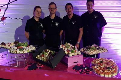Traiteur L'Atelier de Fred La Chapelle-d'Armentières #3 Quatre personnes posent derrière une table garnie de plateaux de bouchées et de plats variés dans un cadre éclairé en violet.