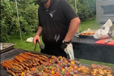 Traiteur L'Atelier de Fred La Chapelle-d'Armentières #5 Homme cuisinant des brochettes et saucisses sur un grand barbecue en plein air, avec un jardin en arrière-plan.