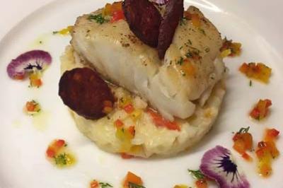 Plat de poisson blanc sur purée, décoré de rondelles de chorizo, légumes colorés et fleurs comestibles.