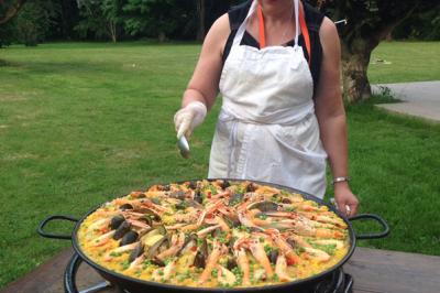 Une femme en tablier prépare une grande paella aux fruits de mer sur un réchaud en plein air, dans un jardin verdoyant.