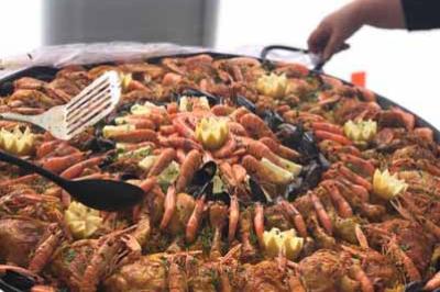 Grande poêle de paella garnie de crevettes, moules, poulet et légumes, avec une main tenant une spatule.