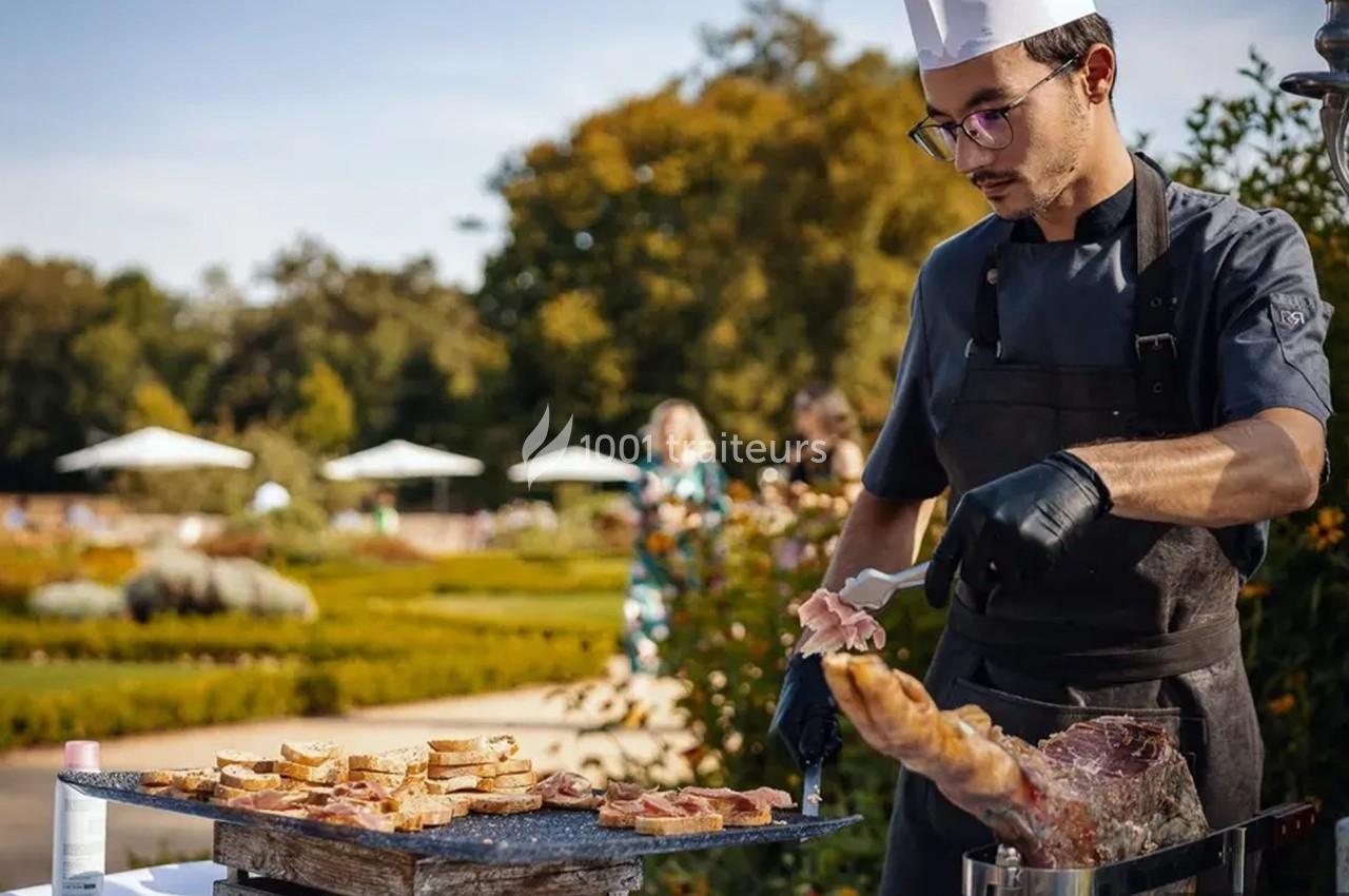 Traiteur Niel Traiteur Le Lion-d'Angers #1 Un chef prépare des tranches de jambon et de pain grillé en extérieur, dans un jardin par une journée ensoleillée.