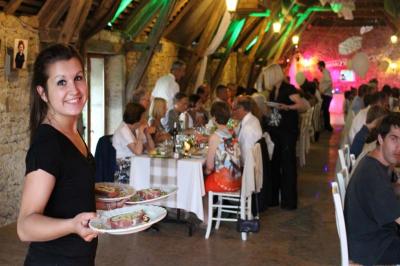 Une serveuse souriante apporte des plats dans une salle de réception rustique avec des convives attablés.