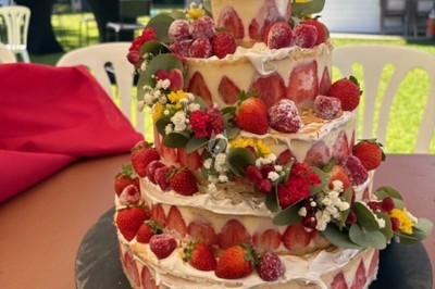 Gâteau à étages décoré de fraises, framboises et fleurs, présenté sur une table en extérieur par temps ensoleillé.
