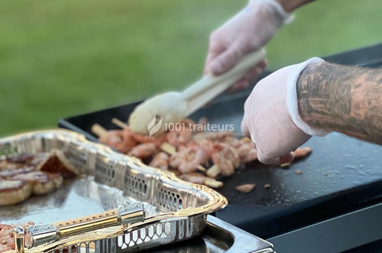 Personne cuisinant des brochettes de crevettes sur une plancha, avec un plateau en métal contenant des aliments à côté.