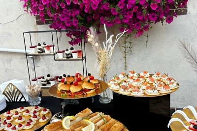 Buffet extérieur avec assortiment de plats, incluant rouleaux frits, mini-burgers et desserts, devant des fleurs violettes.