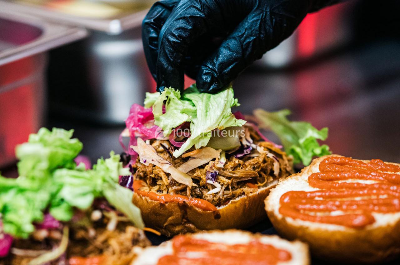 Une main gantée ajoute de la laitue sur un burger garni de viande effilochée, de chou et de sauce.