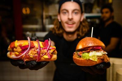 Un cuisinier présente un hot-dog garni et un burger avec des ingrédients frais dans un restaurant.