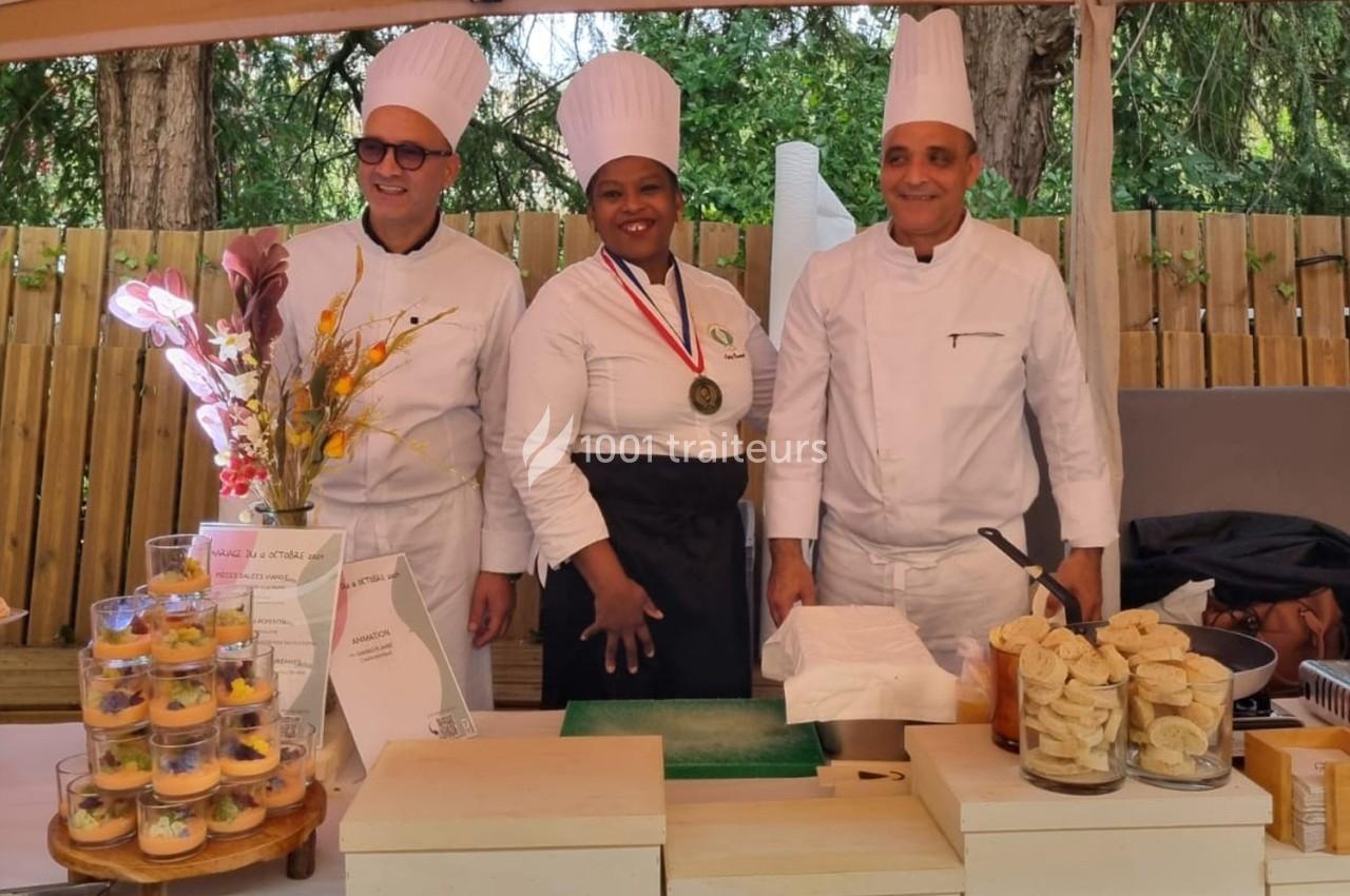 Trois chefs en tenue blanche posent devant une table garnie de verrines, pains et décorations florales, en extérieur.