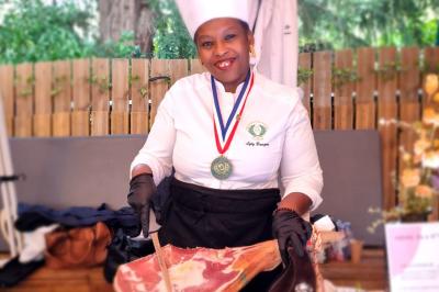 Une cheffe en tenue blanche et toque découpe un jambon sur un stand en extérieur, portant une médaille autour du cou.