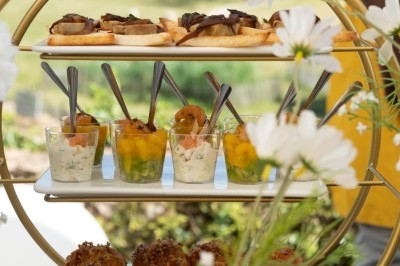 Présentoir doré avec verrines, canapés et petits pains garnis, entouré de fleurs blanches en extérieur verdoyant.