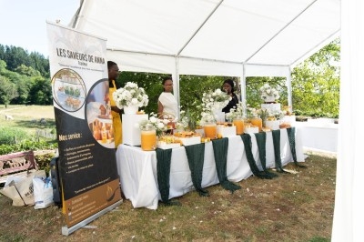 Stand de buffet sous une tente blanche, décoré de fleurs et boissons, avec trois personnes en arrière-plan.