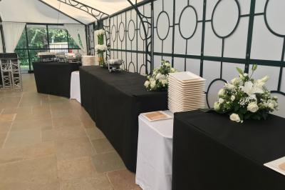 Buffet dressé sous une tente avec nappes noires, assiettes empilées, plats en inox et décorations florales.