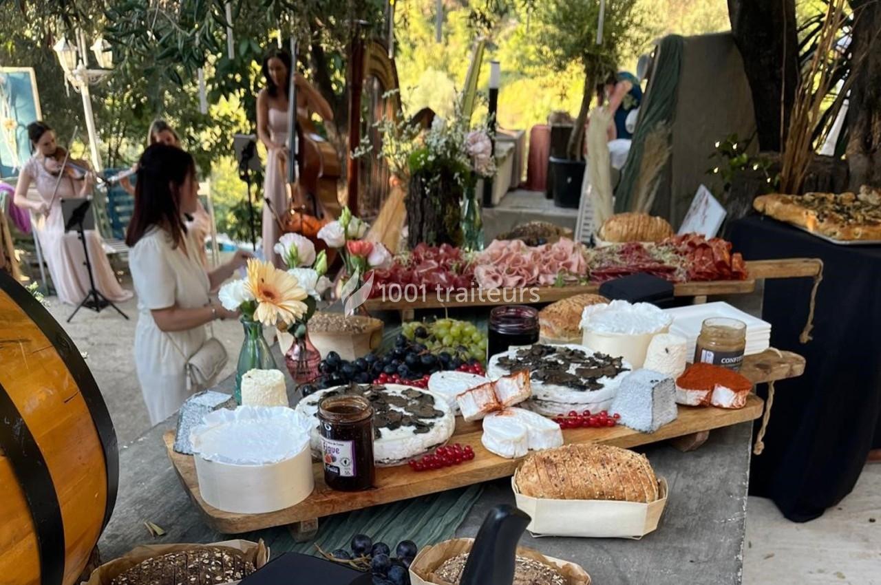 Plateau de fromages, charcuteries et pains sur une table en extérieur, avec des musiciens en arrière-plan.