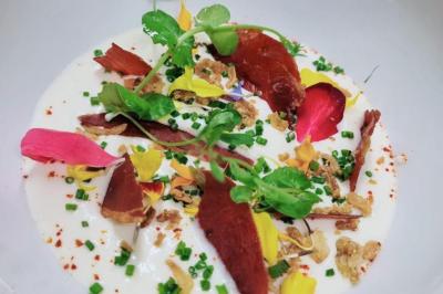 Assiette garnie de crème blanche, pétales de fleurs colorées, herbes fraîches, morceaux de viande séchée et croustillants.