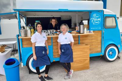 Deux femmes souriantes en tablier devant un food truck bleu clair servant des boissons.