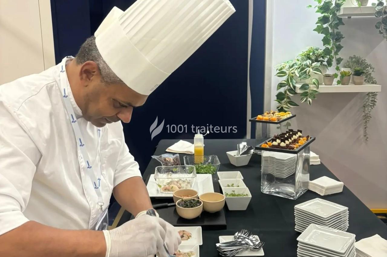Un chef en tenue blanche prépare des plats sur une table noire, avec des assiettes et des ingrédients disposés autour.