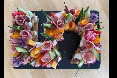 Plateau en forme de couronne garni de charcuterie, légumes frais, fromage et fleurs comestibles sur ardoise noire.
