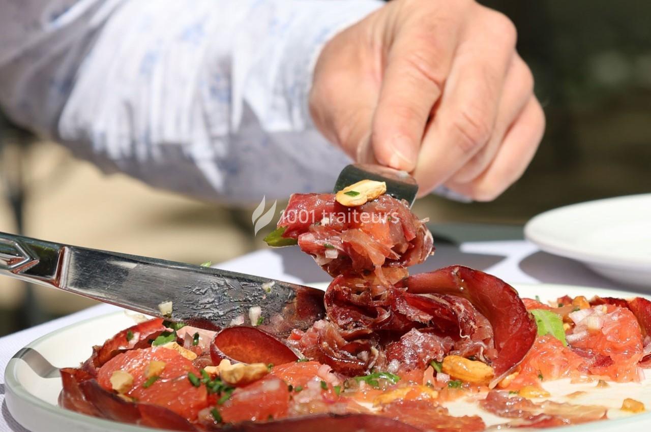 Traiteur Restaurant la Touloubre La Barben #1 Une main utilise un couteau pour servir des tranches de viande séchée garnies de noix et d'herbes sur une assiette.