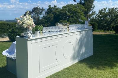 Traiteur Boeuf Traiteur Nice #3 Bar extérieur blanc sur une pelouse, décoré de fleurs et de verres, entouré d'arbres sous un ciel ensoleillé.