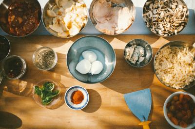 Traiteur Pizzaiolo Tsogani Paris #3 Ingrédients variés disposés sur une table en bois, comprenant œufs, légumes, épices, herbes et fromages.