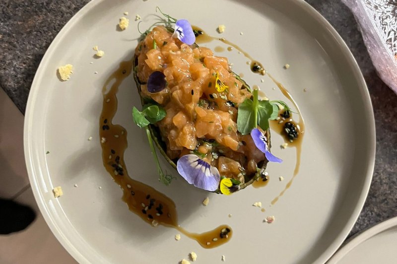 Tartare de saumon présenté dans une coque d'avocat, décoré de fleurs comestibles et entouré de sauce.