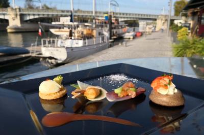 Traiteur Le Lonjoie Saint-Benoît #1 Assortiment de bouchées gastronomiques sur une assiette noire, avec un quai et des bateaux en arrière-plan.