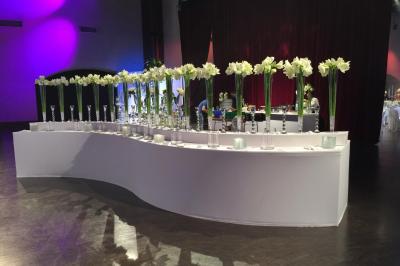 Alignement de vases transparents contenant des fleurs blanches sur une table blanche courbée dans une salle événementielle.