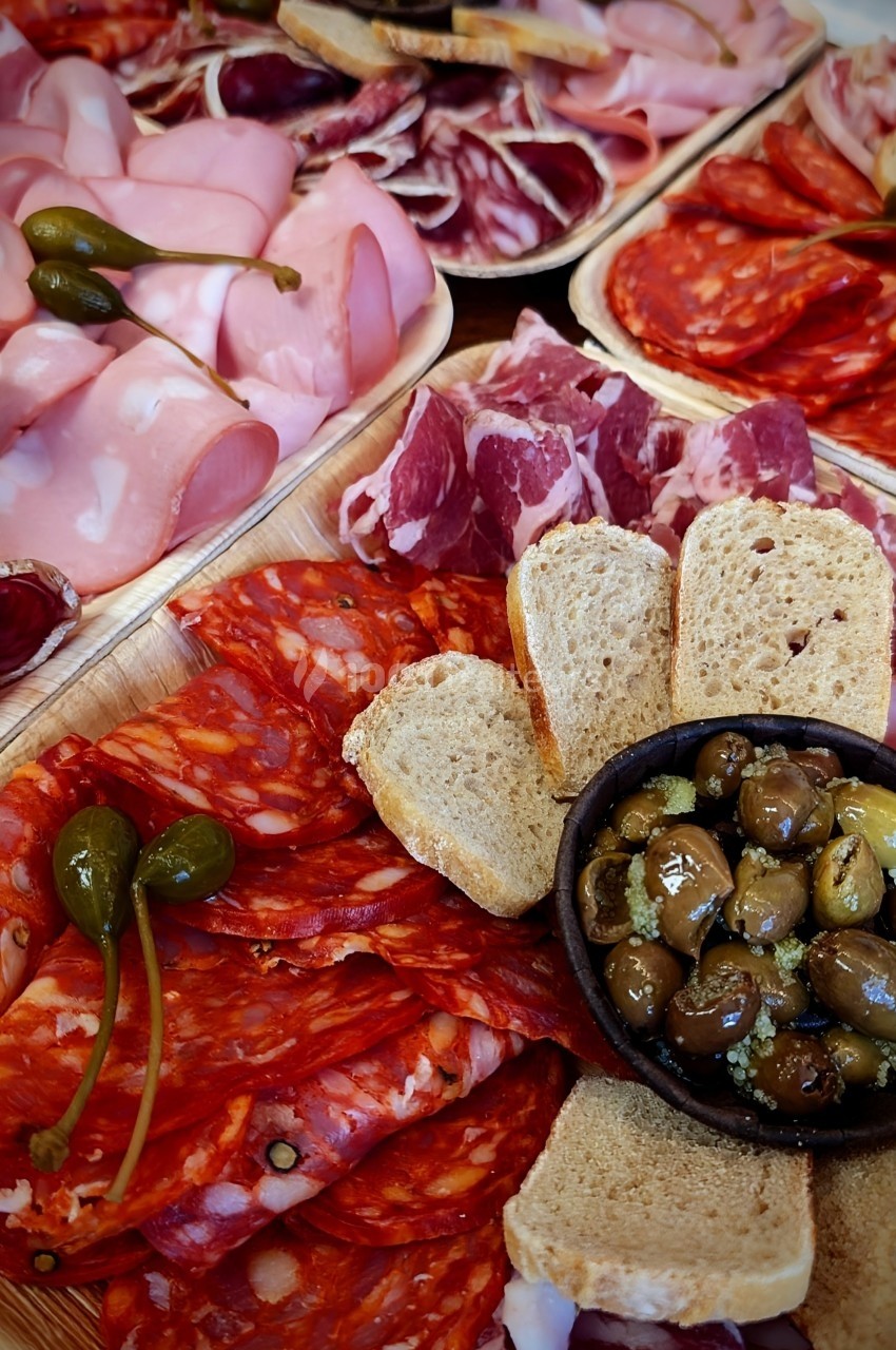 Tagliere di salumi / ©Gusto e cucina Plateau de charcuterie variée avec olives marinées, cornichons et tranches de pain disposés sur un plateau en bois.