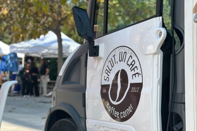 Petit camion de café avec une porte ouverte, stationné dans un espace extérieur entouré d'arbres et de stands.