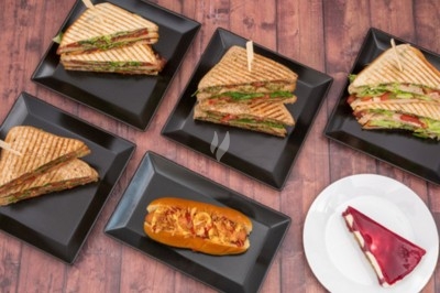 Assortiment de sandwichs grillés sur des assiettes noires et part de gâteau sur une assiette blanche, sur table en bois.