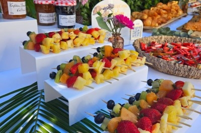 Brochettes de fruits frais disposées sur des supports blancs, avec confitures et viennoiseries en arrière-plan.