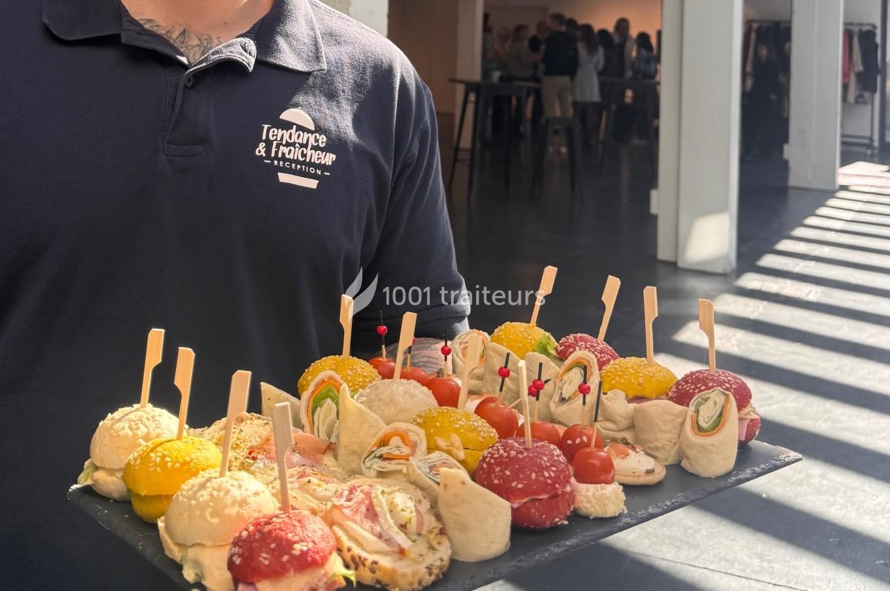 Plateau de bouchées apéritives variées présentées par un serveur dans un espace lumineux avec des invités en arrière-plan.