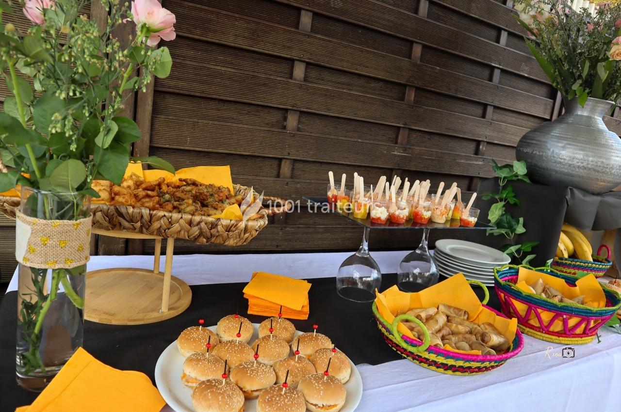 Traiteur Iles en Fete  Magny-les-Hameaux #1 Buffet coloré avec mini-burgers, amuse-bouches, fleurs et paniers garnis, disposé sur une table en extérieur.