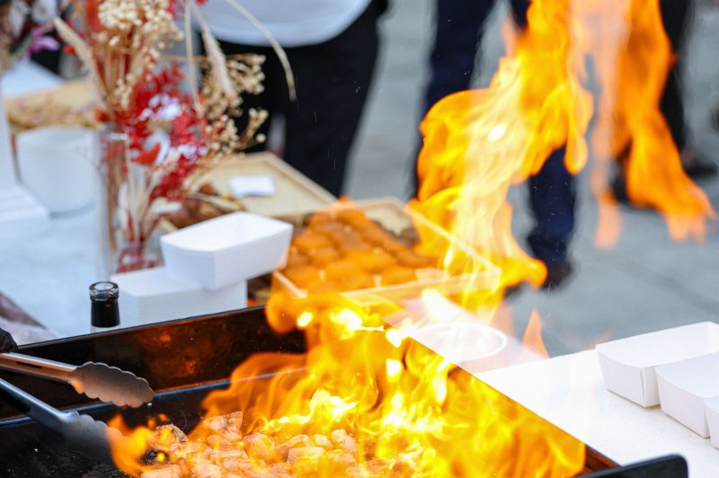 Animation Culinaire Viande grillée en cuisson sur une plancha avec des flammes vives, entourée de décorations florales et de convives en arrière…