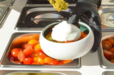 Bol contenant des tomates cerises et une boule de mozzarella, avec une cuillère de pesto ajoutée par une main gantée.