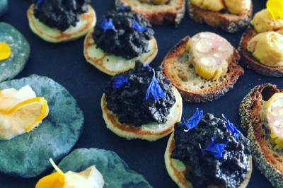 Traiteur Adquat Traiteur Nousty #4 Assortiment de canapés garnis de tapenade, fleurs comestibles et autres ingrédients sur une table sombre.