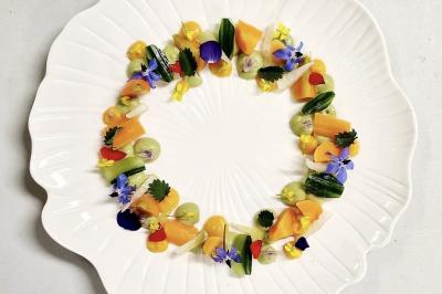 Traiteur Adquat Traiteur Nousty #2 Assiette blanche décorée de légumes colorés, fleurs comestibles et herbes disposés en cercle.
