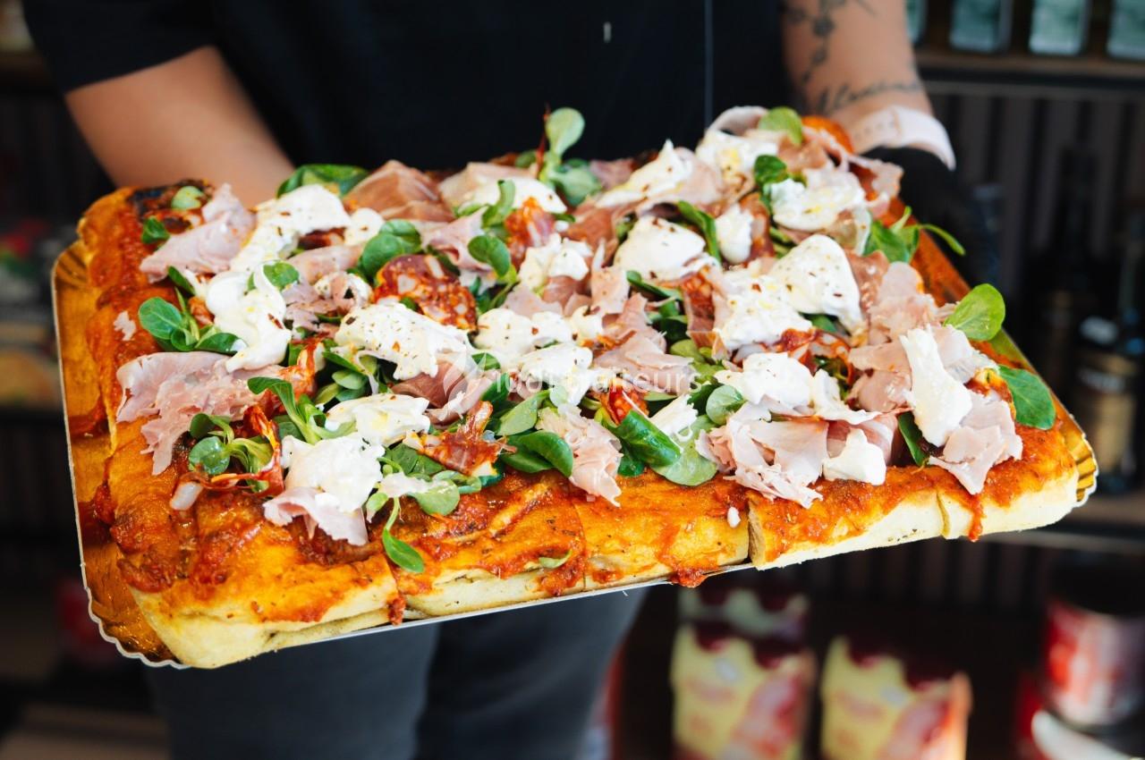 Traiteur Pasta Da Nonna Mia Bouc-Bel-Air #1 Pizza rectangulaire garnie de jambon, fromage, roquette et tomates séchées, tenue par une personne en arrière-plan flou.