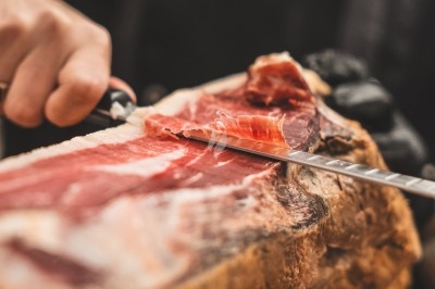 Une main découpe finement une tranche de jambon cru sur un morceau entier avec un couteau.