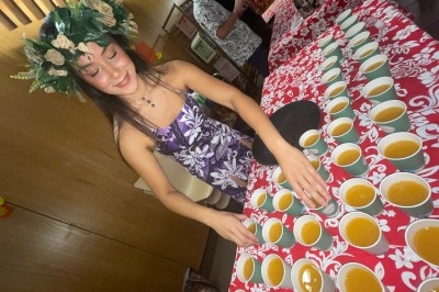 Traiteur Matahiapo Food Le Fuilet #1 Femme en robe fleurie et couronne de feuillage disposant des tasses de boisson sur une table décorée de motifs rouges.