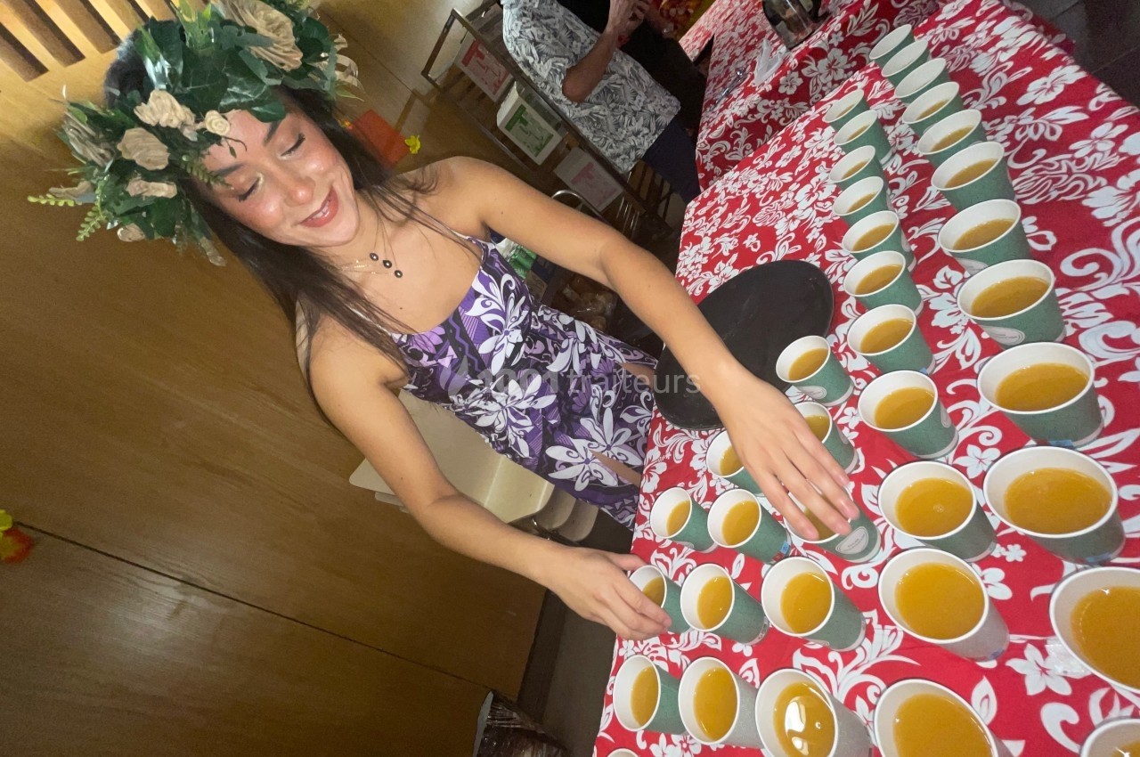 Traiteur Matahiapo Food Le Fuilet #1 Femme en robe fleurie et couronne de feuillage disposant des tasses de boisson sur une table décorée de motifs rouges.